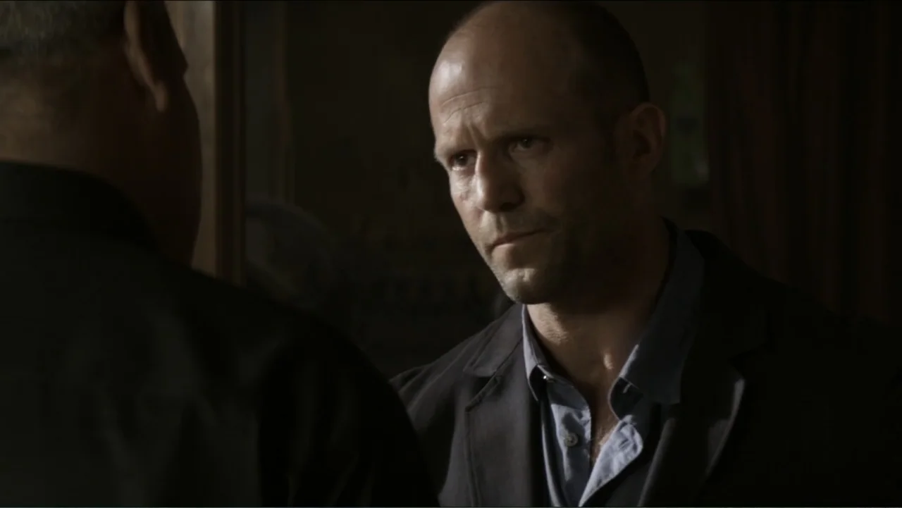 Parker (2013) movie still: medium — A dark, dimly lit interior space with shadows obscuring most architectural details. A bald man wearing a blue…