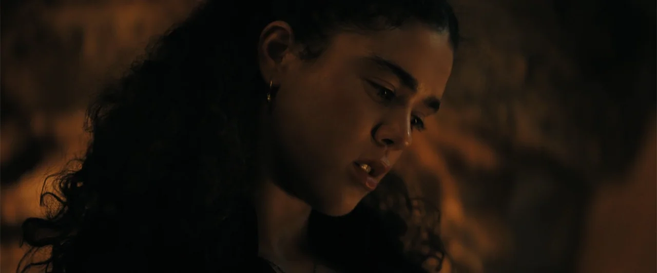 The Bluff (2026) movie still: closeup — A dimly lit cave interior features uneven rock walls and warm, concentrated light sources. A woman with long…