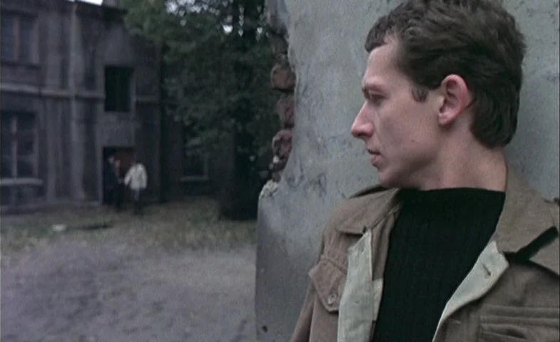 Blind Chance (1987) movie still: medium — A man stands in the foreground next to a crumbling concrete wall. In the background, two figures walk…