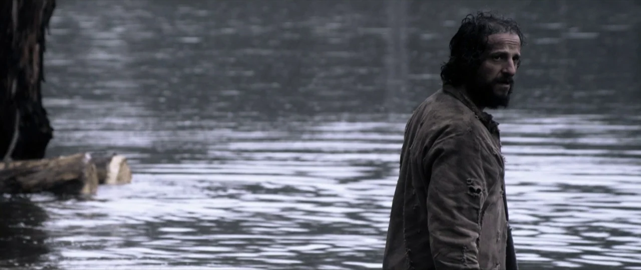 Van Diemens Land (2009) movie still: medium — A man stands chest-deep in a calm, dark-water lake under a cloudy sky. The man wears a tattered brown…