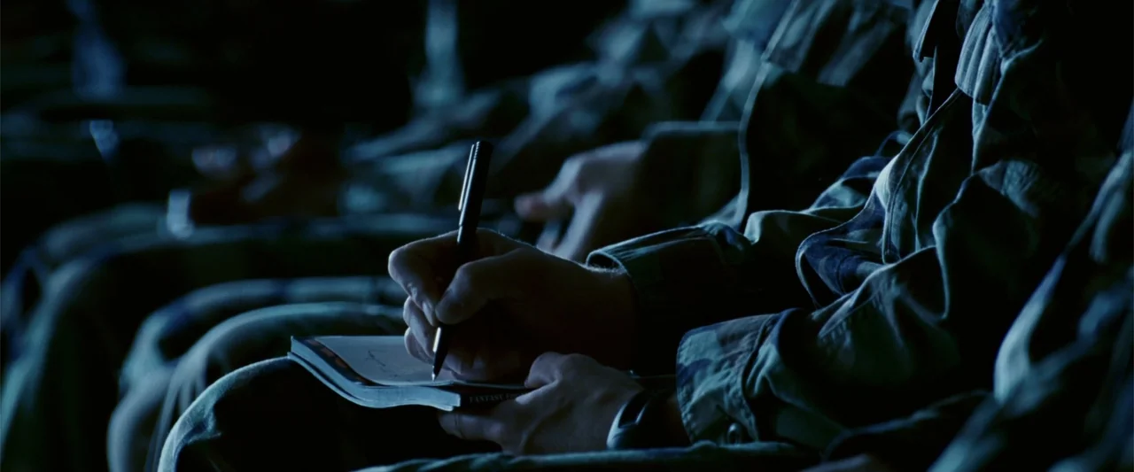 Dear John (2010) movie still: closeup — A dark, dimly lit interior room contains several figures in camouflage uniforms. One individual writes in a…