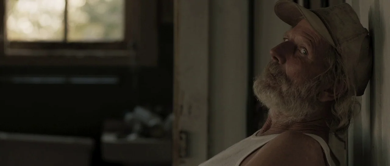 Joe (2013) movie still: medium — An interior space with a window casting light on a textured wall. An elderly man with a beard and baseball cap…