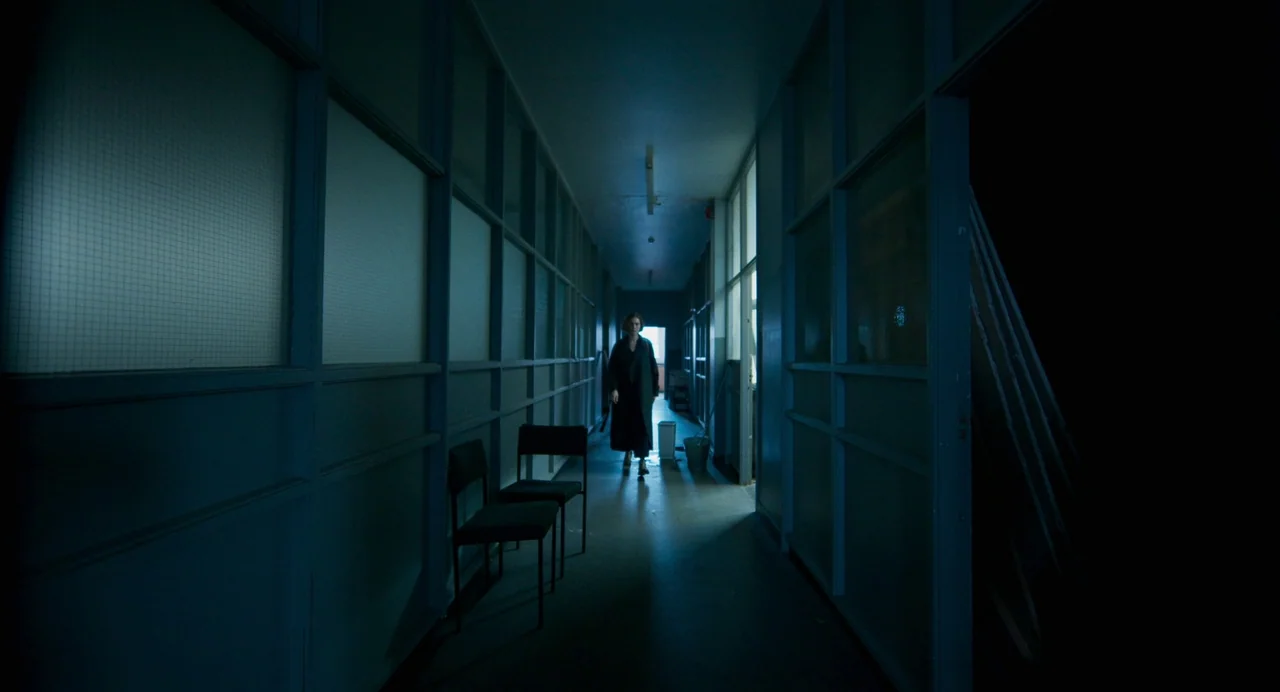 All You Need Is Death (2023) movie still: wide — A long, dimly lit institutional corridor features glass-partitioned offices along both sides. A…