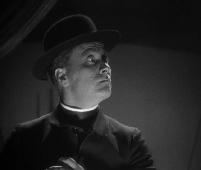 The Threepenny Opera (1931) movie still: medium — A man in a clerical shirt and bowler hat stands in a dark space. He glances upward with a strained…