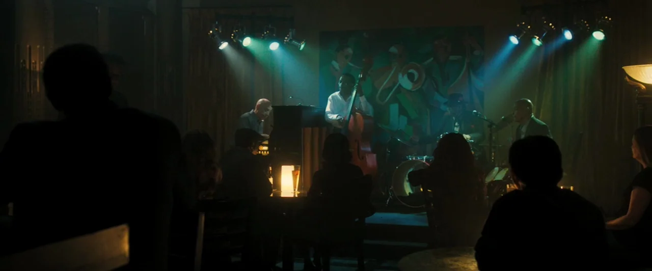 Whiplash (2014) movie still: wide — A dark, dimly lit venue features a small jazz band playing on a stage. Audience members sit at tables in the…