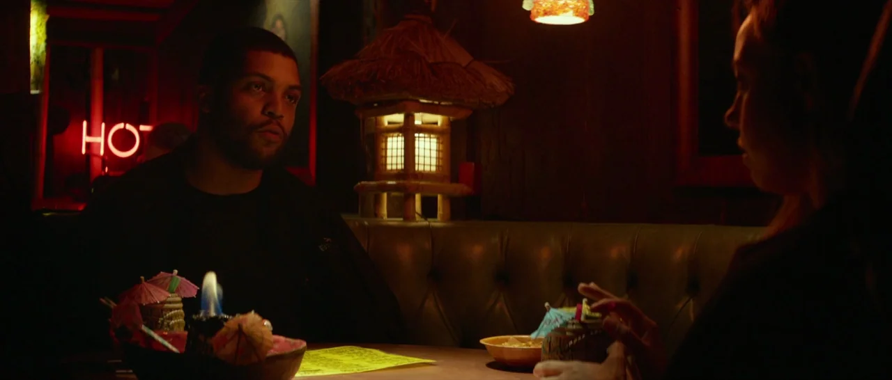 Ingrid Goes West (2017) movie still: medium — A dimly lit tiki-themed restaurant booth features wooden walls and a bamboo-style lamp. A man and…