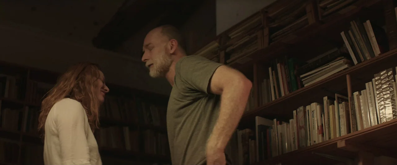 The Only One (2021) movie still: medium — A dimly lit home library features floor-to-ceiling wooden shelves packed with various books. A man and a…