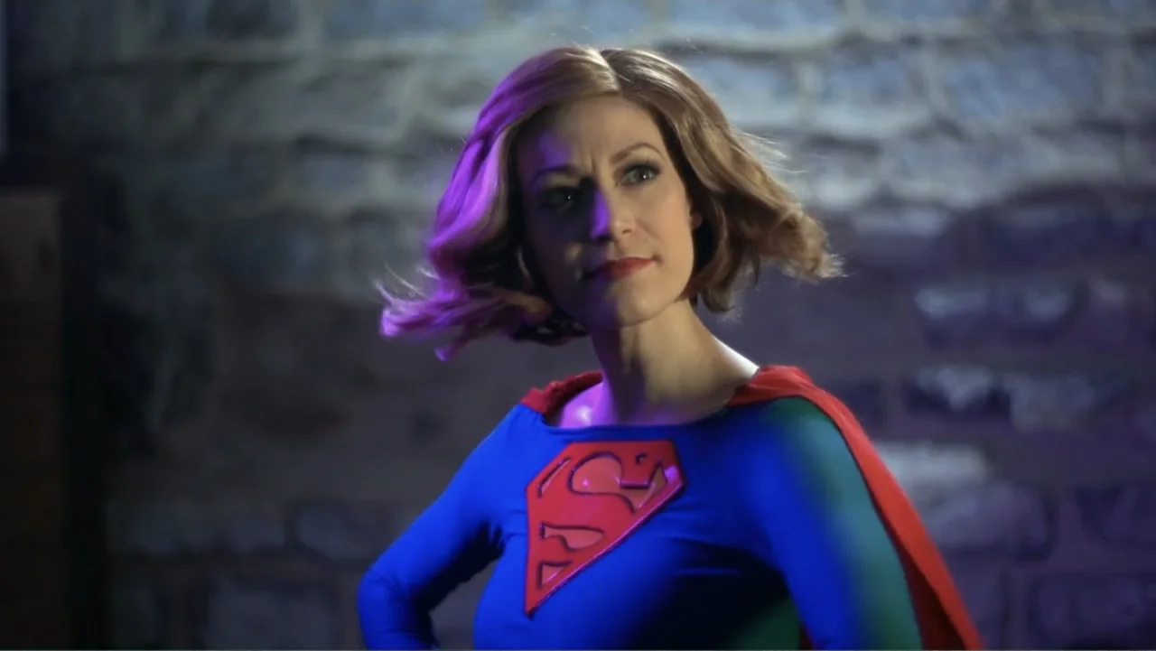 Reign of the Superwomen (2021) movie still: medium — A woman in a blue superhero costume stands against a rough stone wall. She gazes slightly…