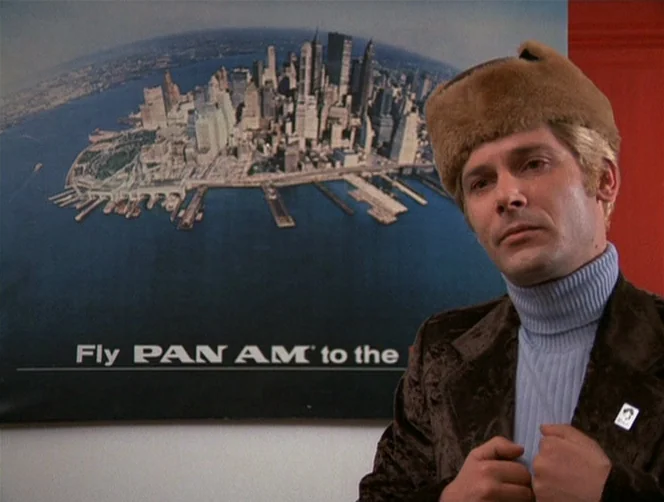 WR: Mysteries of the Organism (1971) movie still: medium — A man stands in an office with a large Pan Am New York poster. He looks forward while…