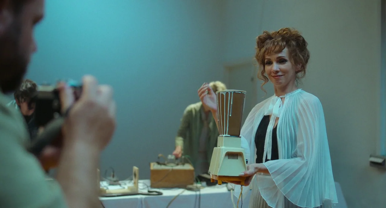 Flux Gourmet (2022) movie still: medium — A stark white laboratory room contains technical equipment and electronic components on tables. A woman in…