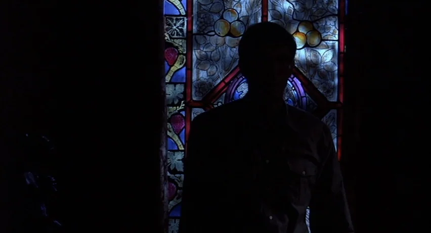Psycho 2 (1983) movie still: medium — A man stands in a dark corridor in front of an ornate stained glass window. The colored light from the window…