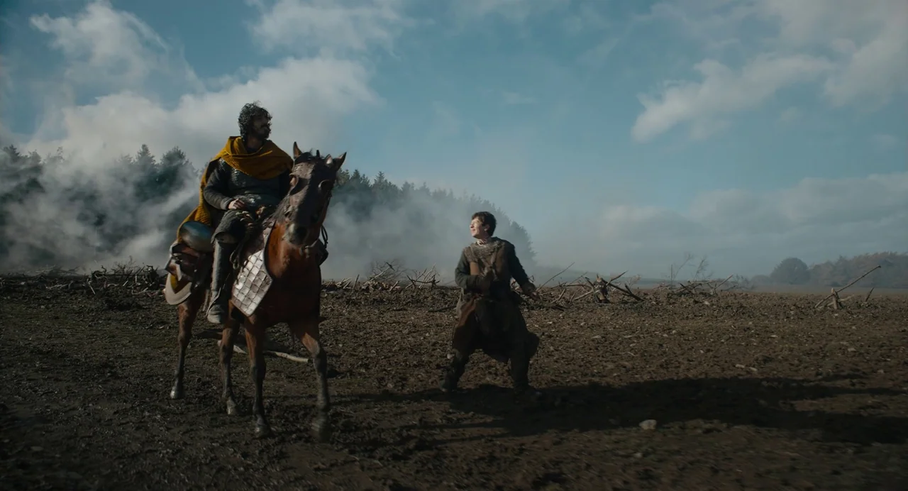 The Green Knight (2021) movie still: wide — A vast, muddy, ploughed field lies beneath a partially cloudy sky. A man rides a horse while his…
