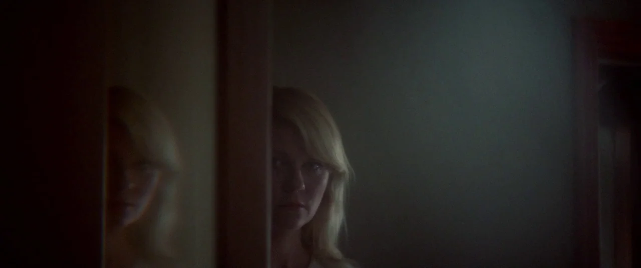 Woodshock (2017) movie still: closeup — A dark, narrow corridor features a partially open doorway obscuring part of the view. A woman with blonde…