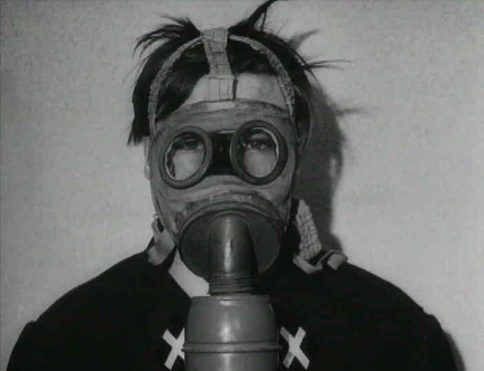 Les Carabiniers (1963) movie still: closeup — A person stands directly before a flat wall in a sterile environment. They wear a vintage rubber gas…