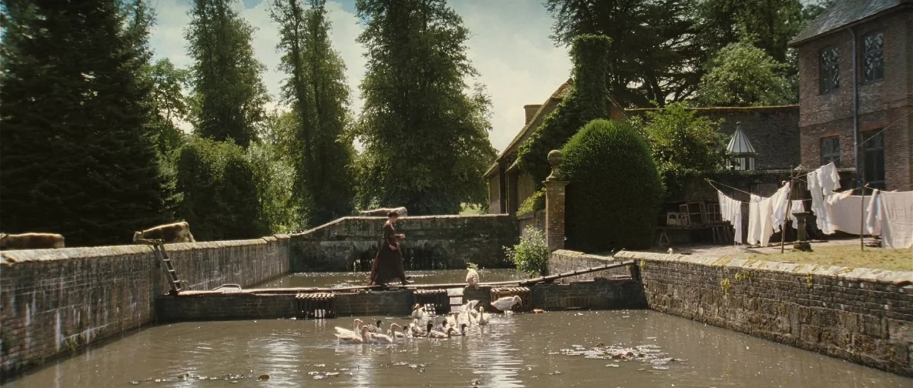 Pride & Prejudice (2005) movie still: wide — A rural English estate features a stone-walled pond and a brick outbuilding nearby. A woman in a period…