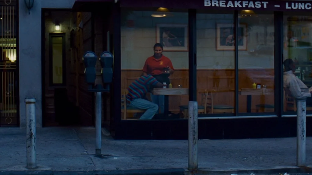 Sugar (2008) movie still: wide — A sidewalk street corner features a storefront window looking into a lit restaurant interior. A server holds a…