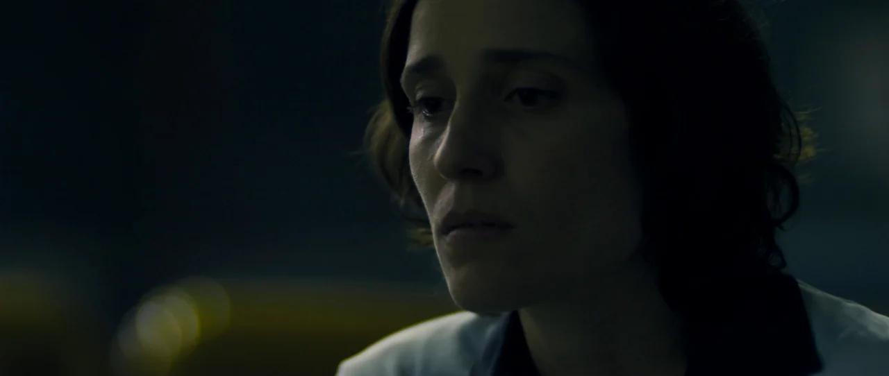 Alps (2011) movie still: closeup — A dimly lit interior space with a dark background and soft yellow light reflections. A woman looks downward with…
