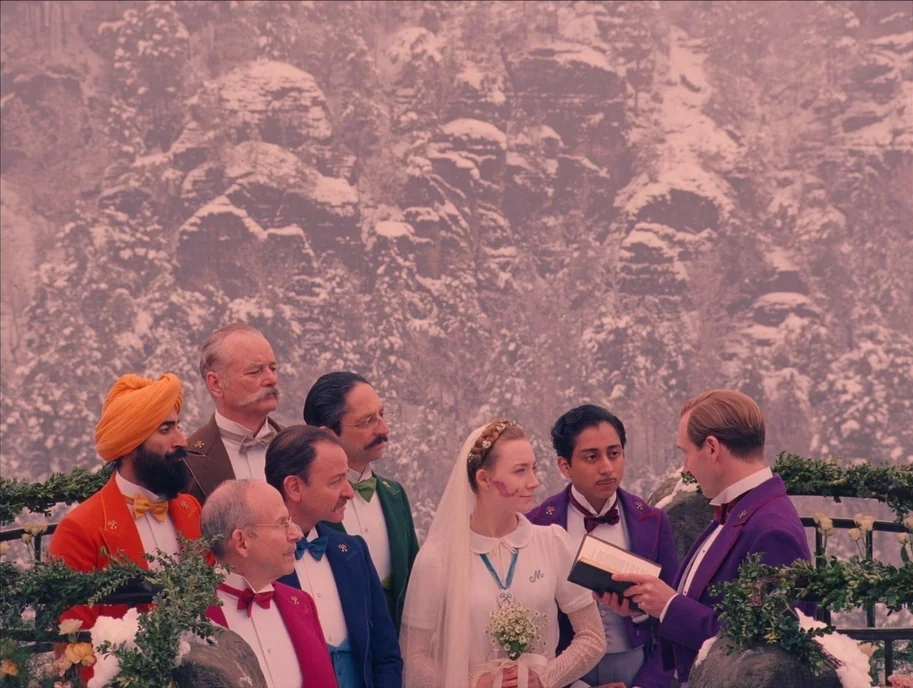 The Grand Budapest Hotel (2014) movie still: medium — A snowy mountain ridge provides the backdrop for a wedding ceremony with formal participants.…