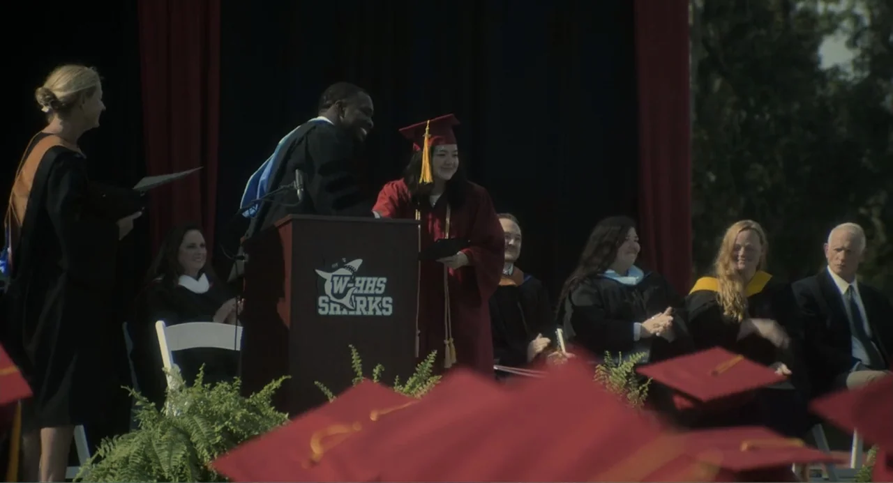 May December (2023) movie still: medium — A graduation ceremony takes place outdoors with faculty members seated on a stage. A graduating student…