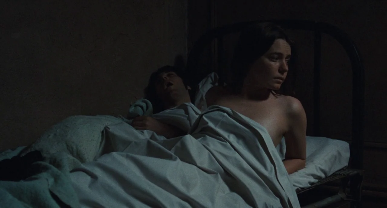 The Panic in Needle Park (1971) movie still: medium — A dark, spartan room features a metal frame bed covered in white linens. A woman sits upright…