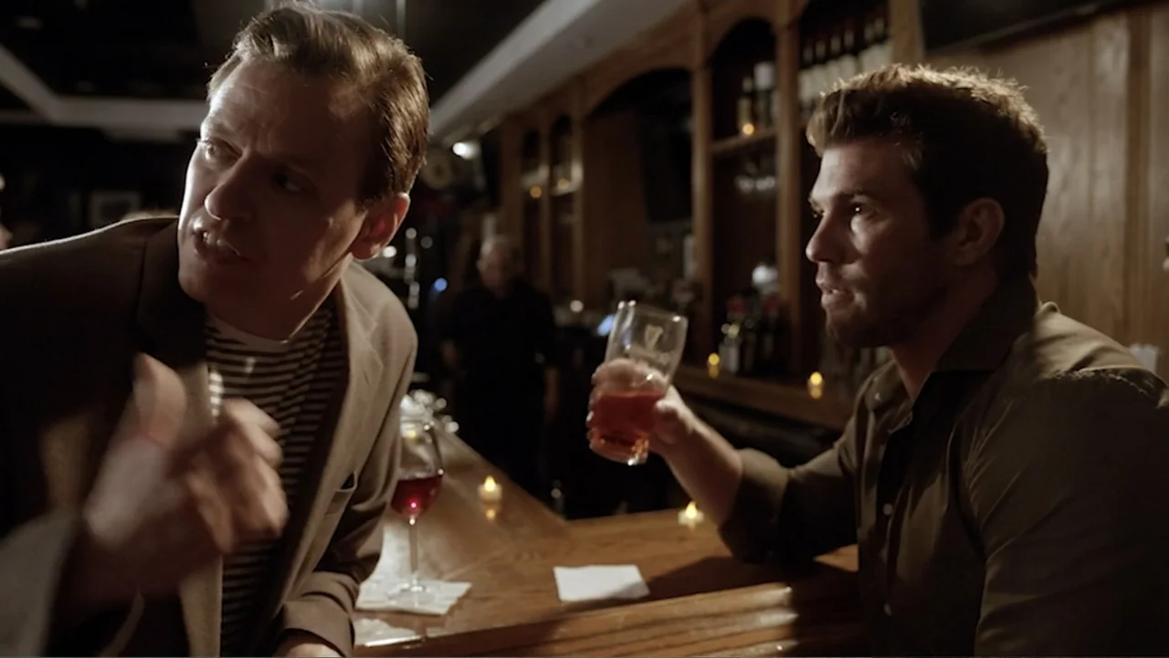 Ask Me to Dance (2022) movie still: medium — A dimly lit bar interior featuring a wooden counter and small glowing candles. Two men sit at the bar…