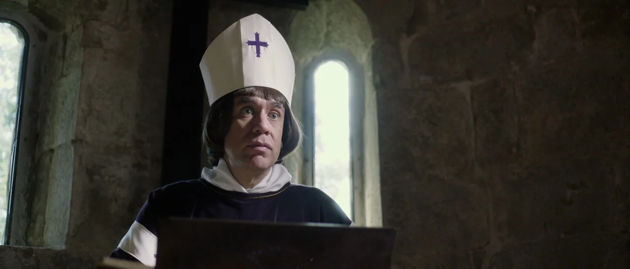 The Little Hours (2017) movie still: medium — A stone room with rugged masonry walls and arched window openings. A man in a miter and ecclesiastical…