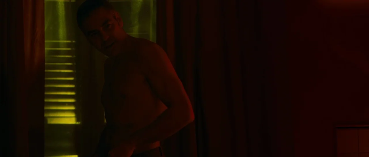 The American (2010) movie still: medium — A dimly lit room features a window with blinds filtering a vibrant green light. A shirtless man stands…