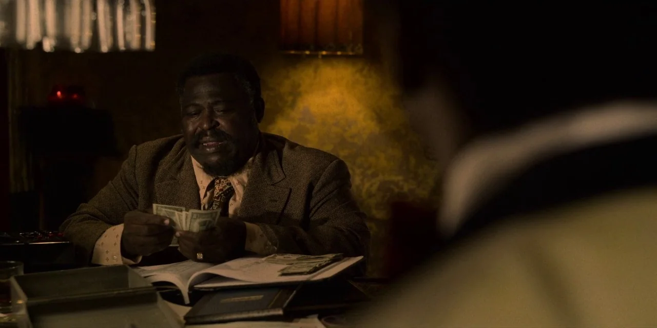 Dolemite Is My Name movie still: medium — A dimly lit, wood-paneled interior office with yellow-hued textured wallpaper behind a man. A black man in…
