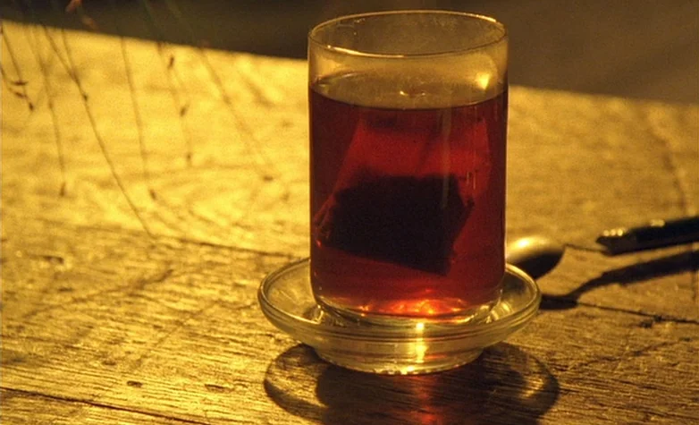 The Double Life of Véronique (1991) movie still: closeup — A glass of tea with a visible teabag rests on a textured wooden table outdoors. A silver…