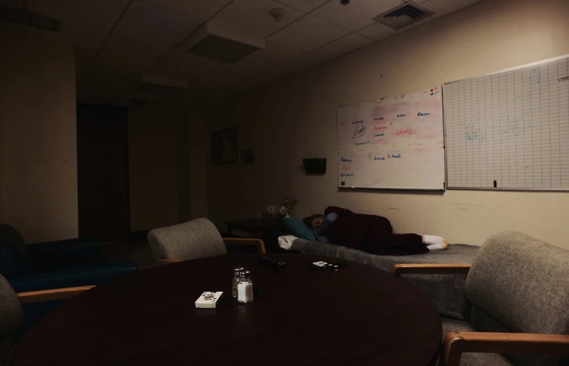 Unsane (2018) movie still: wide — A dimly lit waiting area features a round table, chairs, and a white whiteboard. A person sleeps on a narrow cot…