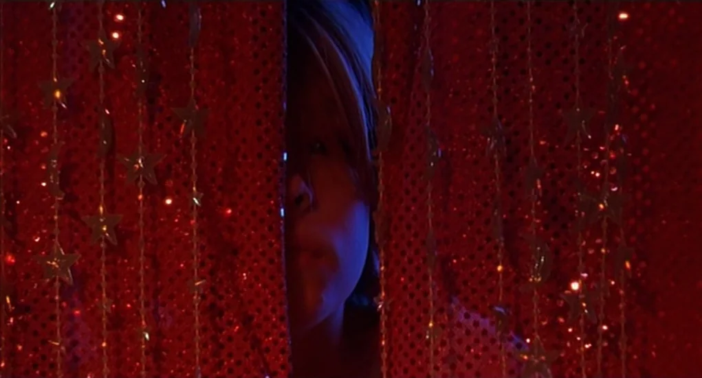 The Heart Is Deceitful Above All Things (2004) movie still: closeup — A person looks through a gap in a dark red beaded star curtain. The red…