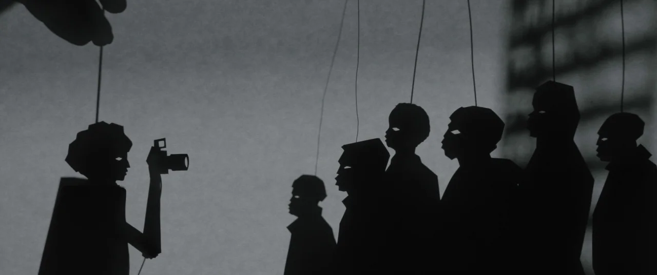 Candyman (2021) movie still: wide — A flat shadow puppet display shows black figures held by strings against a wall. One figure holds a rectangular…