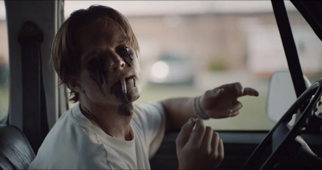 Eye for an Eye (2025) movie still: medium — Inside a parked car, a young man sits in the driver seat during the day. He holds a cigarette and points…