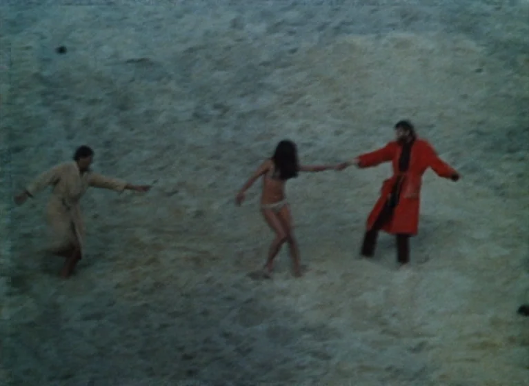 Fruit of Paradise (1970) movie still: wide — A wide shot shows three people standing on a vast expanse of pale sand. The figures walk in a line…