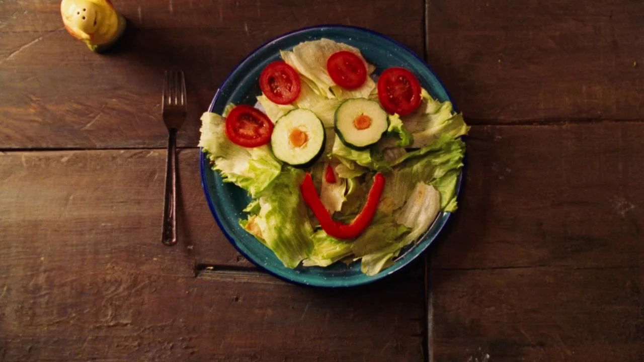 Nacho Libre (2006) movie still: closeup — A dark wooden tabletop holds a blue plate with a salad arranged into a face. A metal fork and a small…