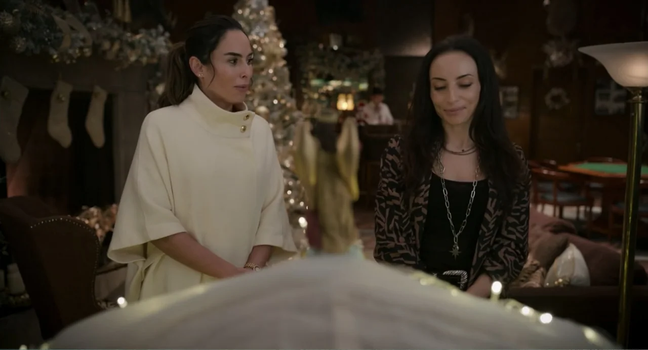 La posada (2023) movie still: medium — A wood-paneled room is decorated for the holidays with a lit Christmas tree in the background. Two women…