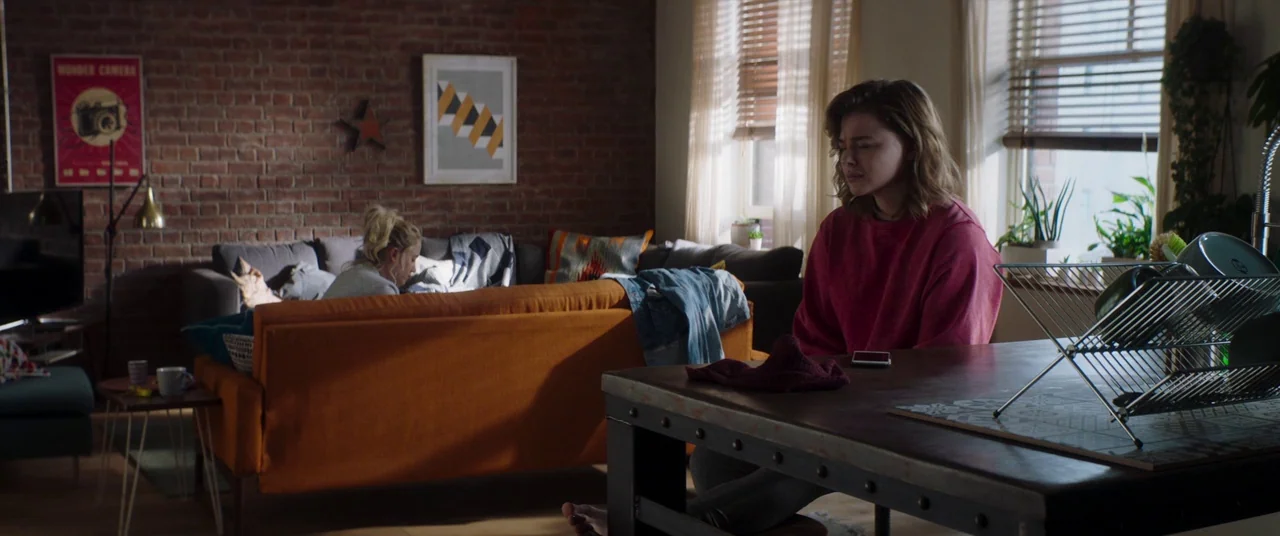 Greta movie still: medium — An apartment interior features a brick wall, orange couch, and a dark wooden table. One woman sits at the table in the…