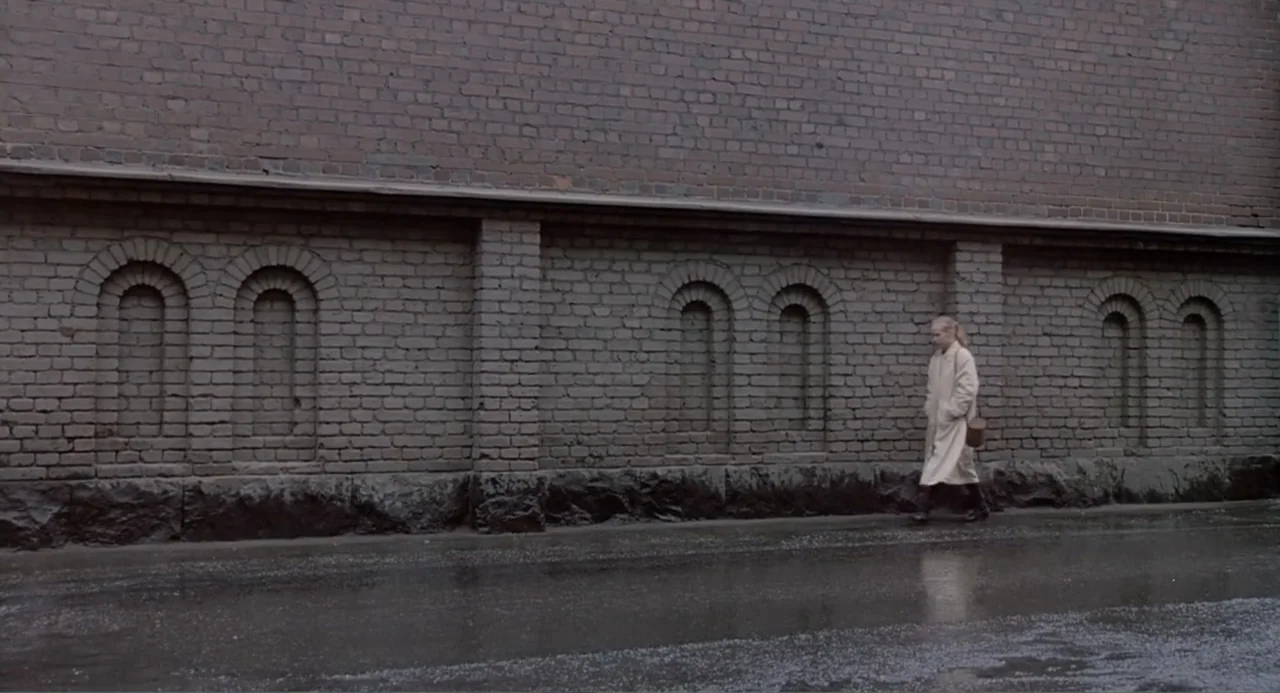 The Match Factory Girl (1990) movie still: wide — A wet street lined with a tall, weathered brick wall featuring recessed arched alcoves. A woman…