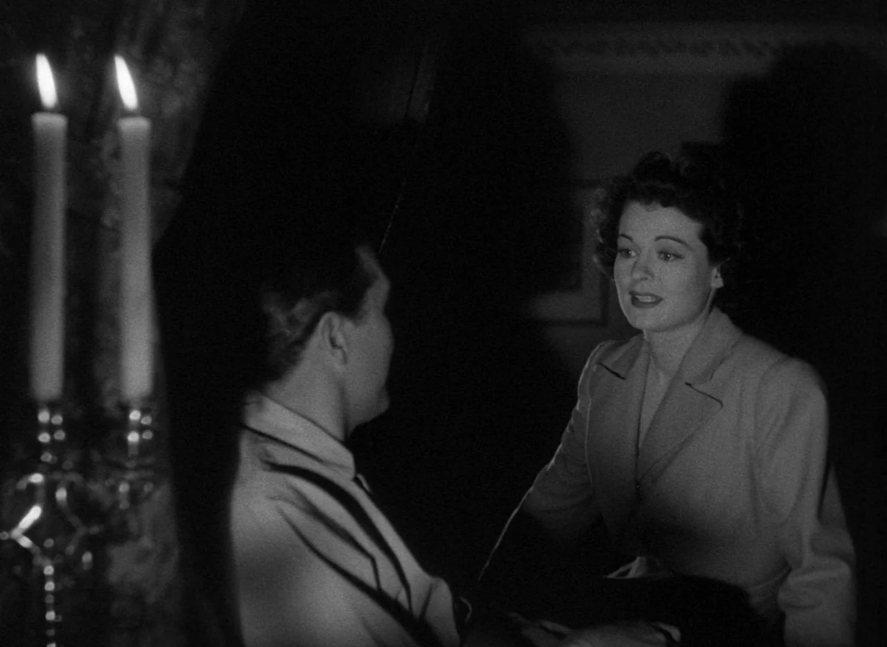 The Uninvited (1944) movie still: medium — A dimly lit room with a man and woman conversing by candlelight. The woman sits facing the camera while…