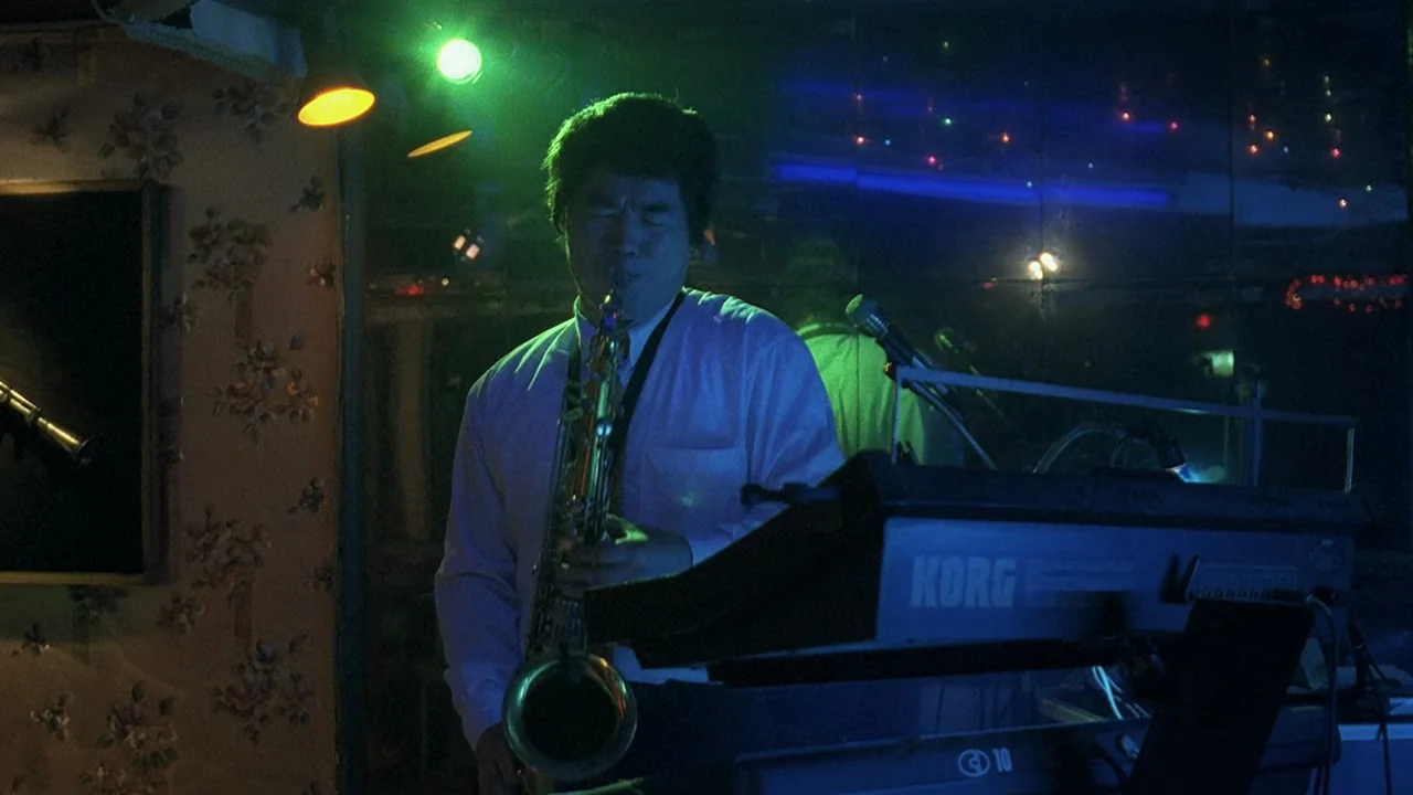 Peppermint Candy (1999) movie still: medium — A dimly lit club interior featuring floral wallpaper and a mirrored wall behind the performer. A man…