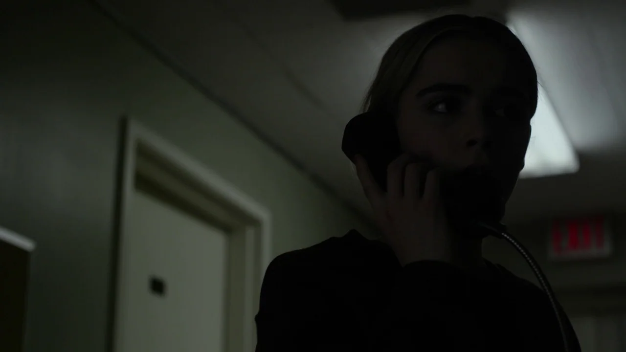 The Blackcoat’s Daughter (2015) movie still: closeup — A dark, dimly lit corridor features a doorway and an illuminated exit sign. A woman in…