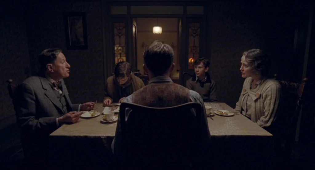 The King’s Speech (2010) movie still: medium — A dark dining room with patterned wallpaper and an overhead pendant light. Four people are seated at…