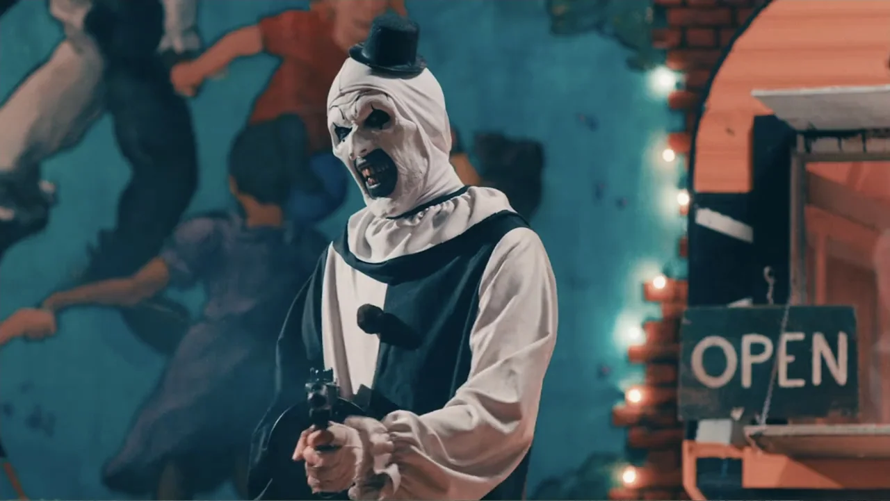 Terrifier 2 (2022) movie still: medium — A dark carnival area features a mural of children and a string of lights. A clown in a black and white…