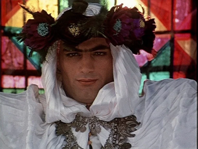 Ashik Kerib (1988) movie still: closeup — A man stands before a geometric stained glass window with multicolored panes. He wears a white draped…