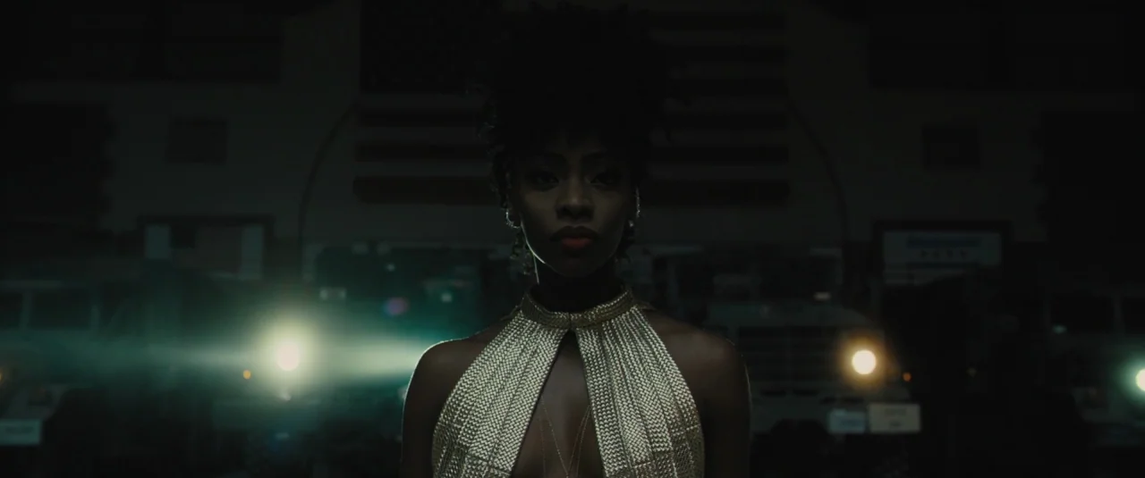 Chi-raq (2015) movie still: medium — A dimly lit interior space with large military-style trucks parked in the background. A woman in a gold dress…