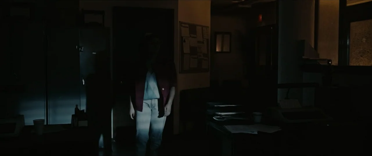 Vicious Fun (2021) movie still: medium — A dimly lit room contains desks with papers and a notice board. A woman stands in the center of the dark…