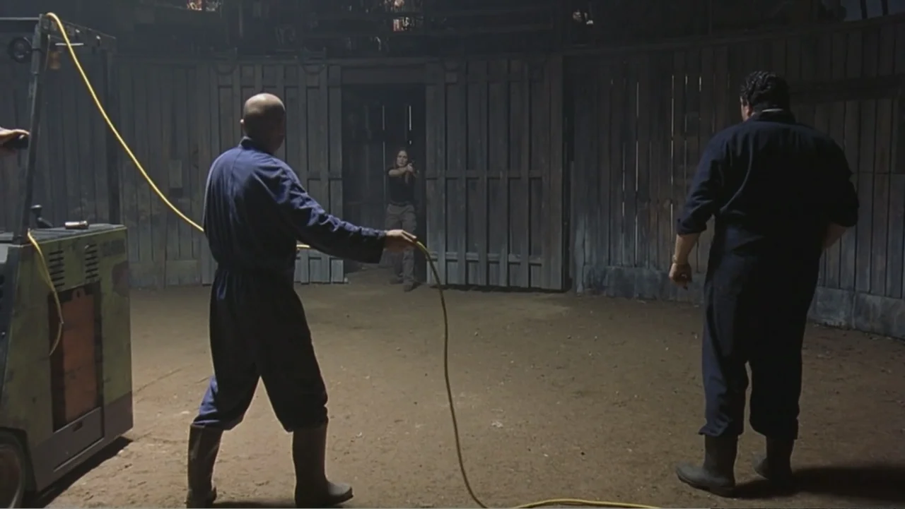Hannibal (2001) movie still: fullbody — A dark wooden structure features a central doorway and dirt floor. Two men stand in front of a woman who…