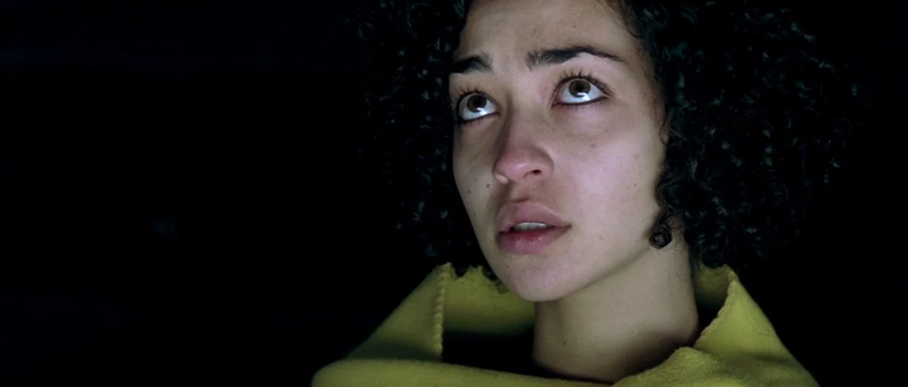 Isolation (2005) movie still: closeup — A person is centered in a pitch-black environment with no visible background details. She looks upwards with…