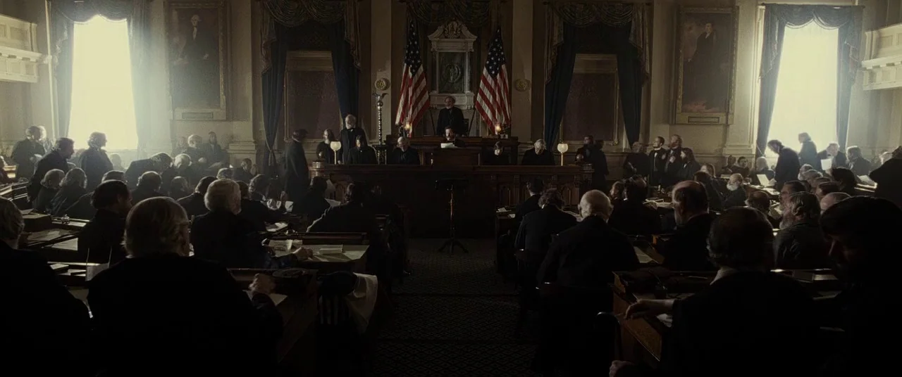 Lincoln (2012) movie still: wide — A dimly lit 19th-century legislative hall features wooden desks and American flags. Numerous men sit in rows…