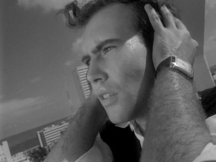 I Am Cuba (1964) movie still: closeup — A rooftop terrace overlooks an urban coastal landscape featuring multi-story buildings and ocean. A young…
